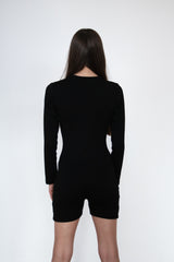 Back view of a woman wearing the cosycore black button up romper from becosy which has shorts, long sleeves, and an embroidered logo