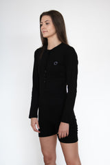 Side view of a woman wearing the cosycore black button up romper from becosy which has shorts, long sleeves, and an embroidered logo