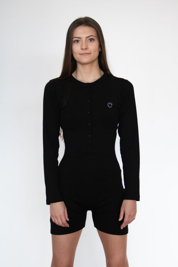 Front view of a woman wearing the cosycore black button up romper from becosy which has shorts, long sleeves, and an embroidered logo