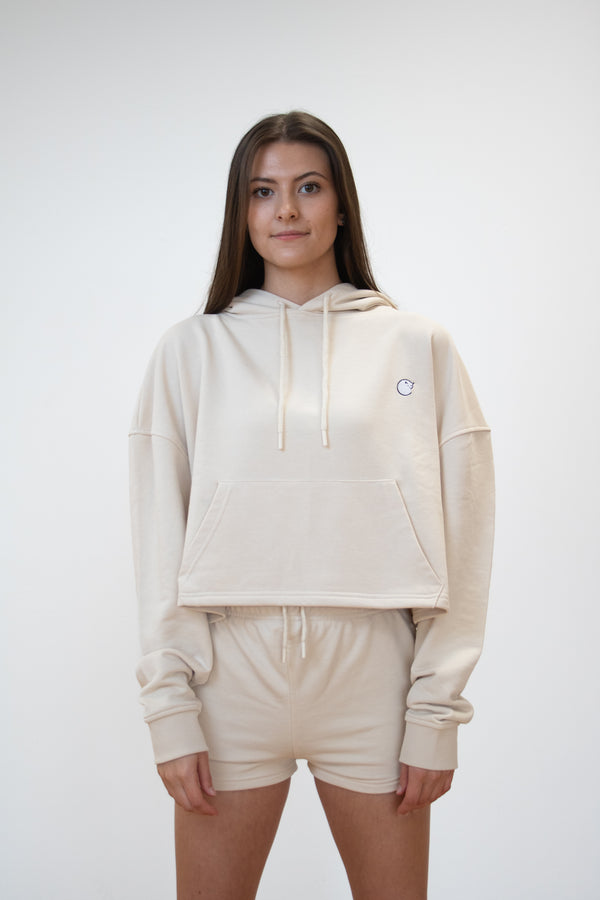 Front view of female model wearing cosycore cropped hoodie in cream showing front pocked and embroidered logo. matched with cosycore terry shorts
